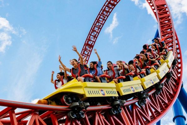A red rollercoaster with people with their arms in the air in front of a blue sky.